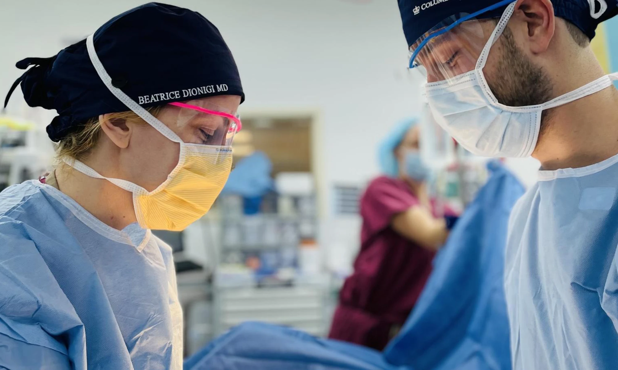 Surgeon Beatrice Dionigi, MD, FACS, FASCRS, wearing surgical scrubs, mask, and face shield, operates alongside another masked surgeon in an operating room.