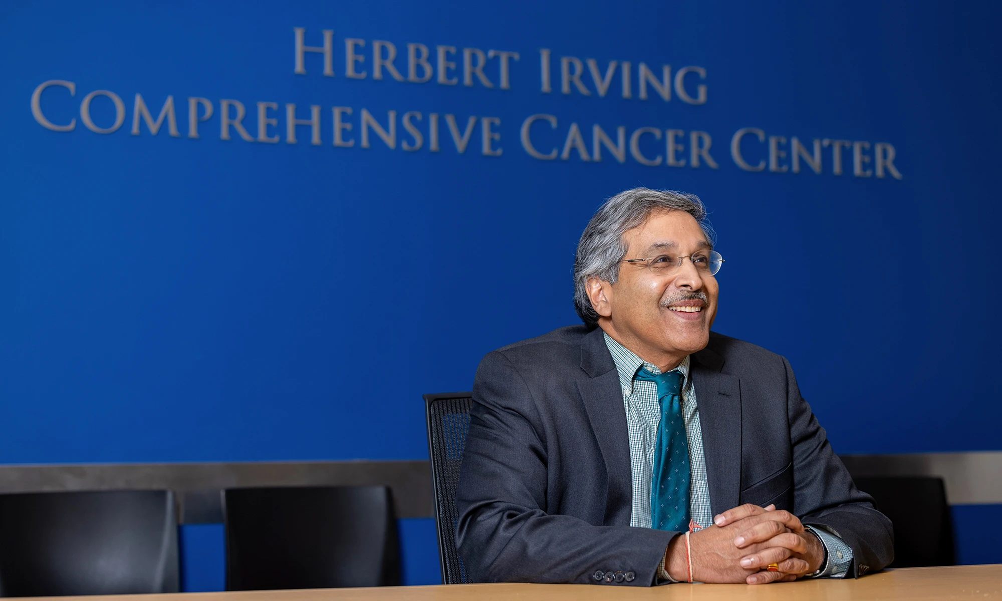 Herbert Irving Comprehensive Cancer Center Director Anil K. Rustgi stands on a staircase against a dark brown background, wearing a suit and blue tie with his hands folded in front of him. 