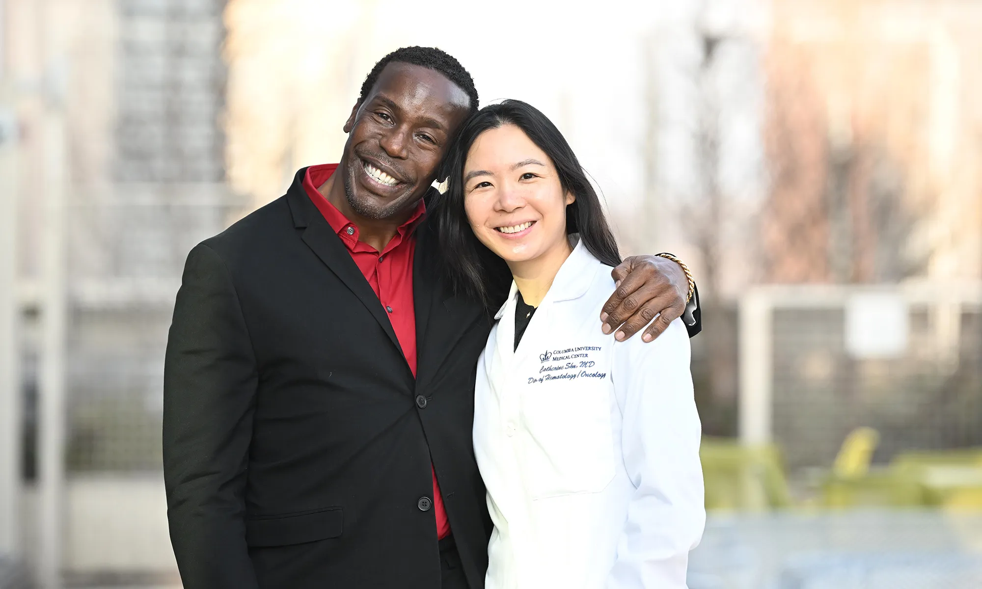 Patient Oswald Peterson stands next to his oncologist Catherine Shu with his arms around her shoulder, both smiling at the camera.