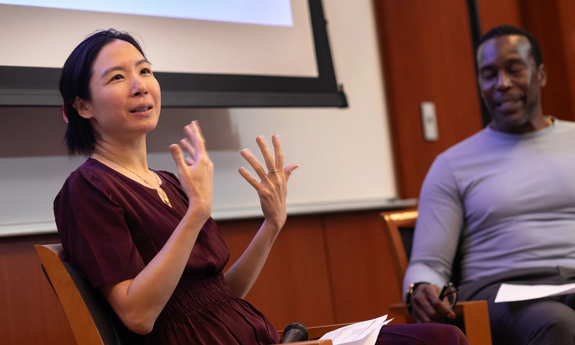 Dr. Catherine Shu speaks while gesturing with her hands during a conversation with her patient Oswald, who sits beside her and listens with a smile. They are seated in front of a projection screen in the Irving Cancer Research Center auditorium.