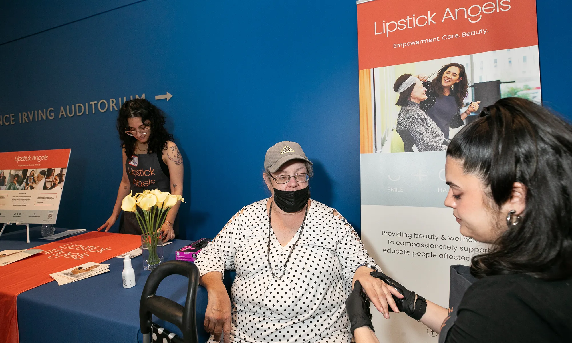 At the Lipstick Angels exhibitor table, a volunteer gives a seated attendee a hand massage, with the group’s banner and materials displayed in the background during the inaugural Cancer Survivors Month event hosted by the Herbert Irving Comprehensive Cancer Center.