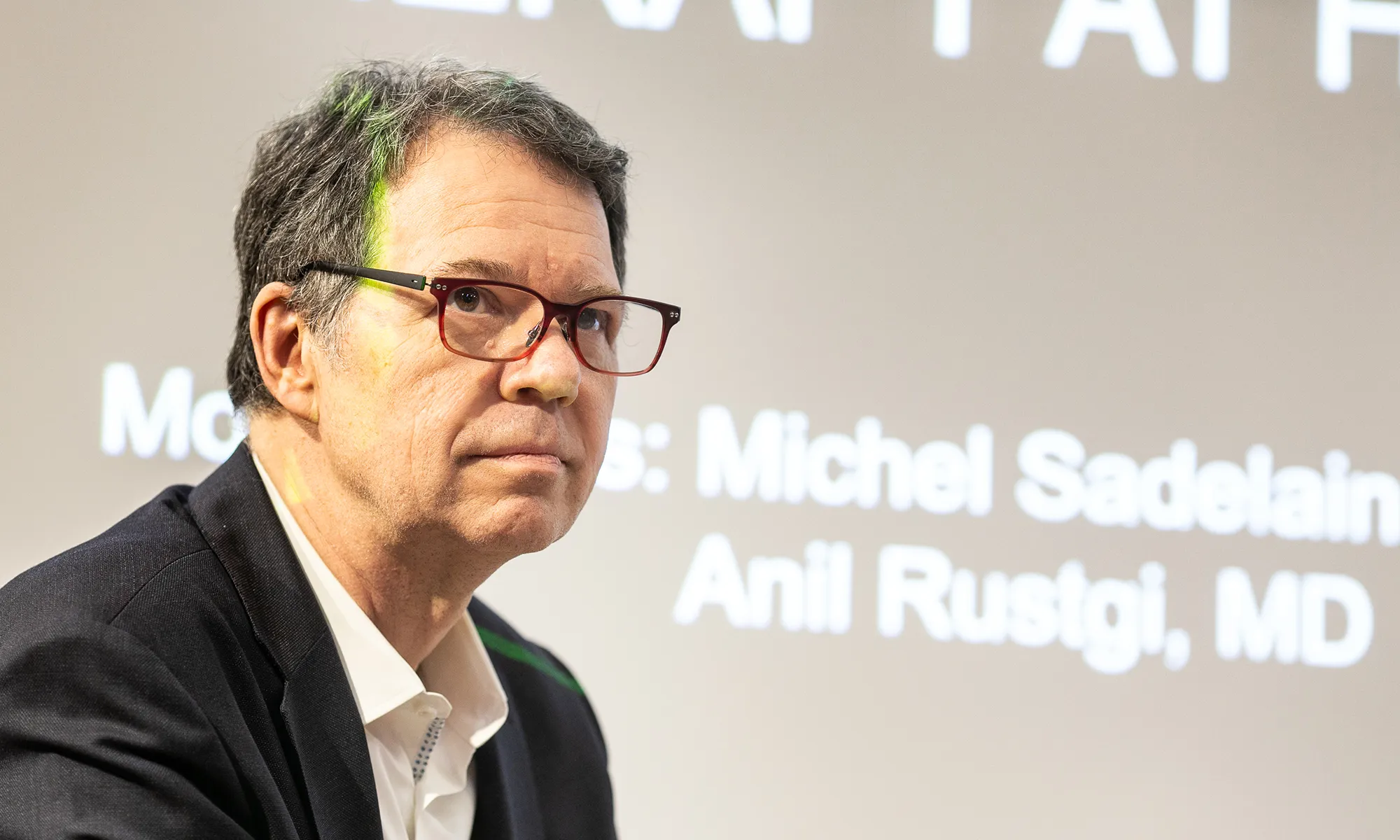 A man wearing glasses and a dark blazer sits in front of a projection screen with blurred text, looking slightly to his left during a presentation or discussion.