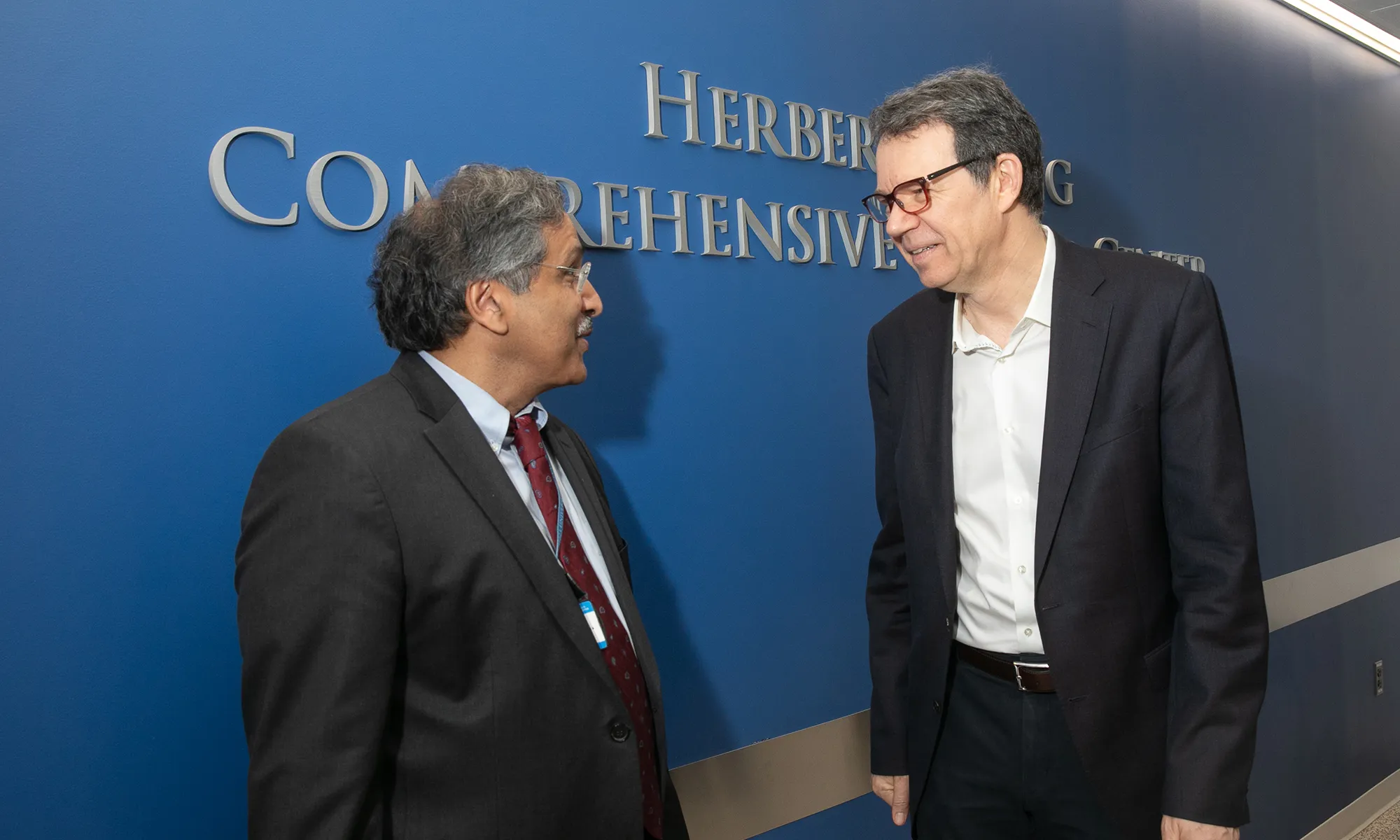 Two men in business attire stand facing each other in conversation against a blue wall with raised lettering in the background.