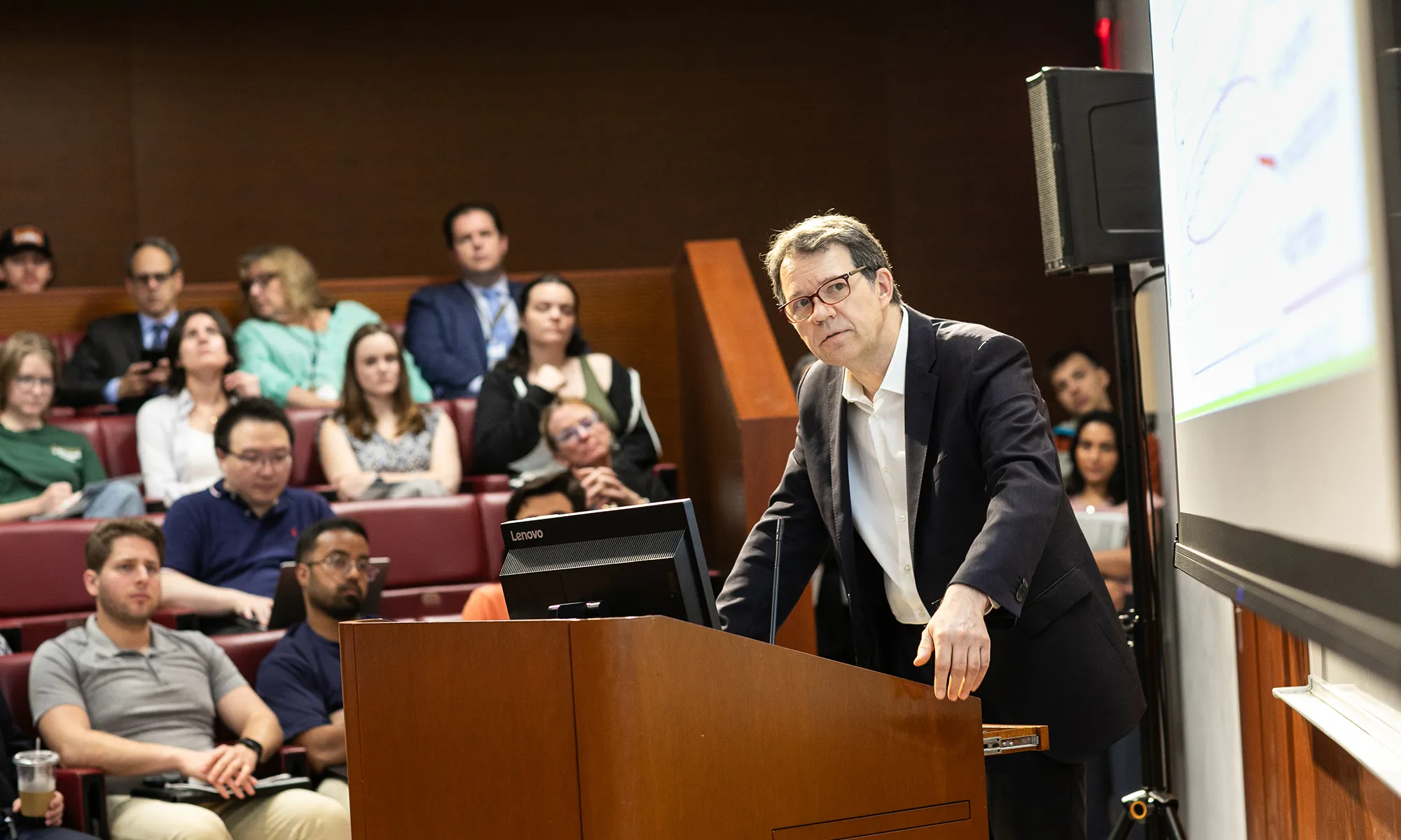 A man in a dark blazer stands at a lectern speaking to an audience in a lecture hall, with attendees seated and listening while a projected slide is visible on the right.