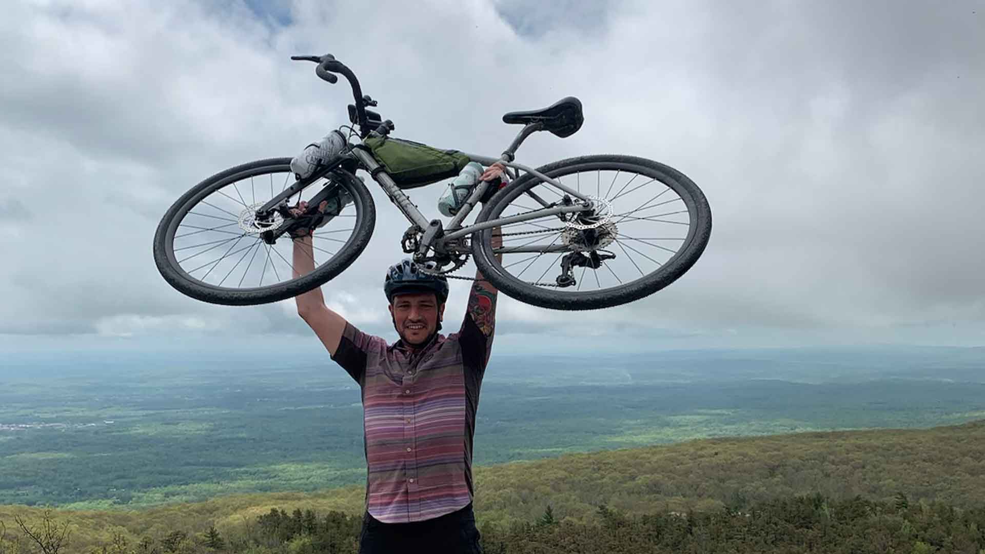 Danny lifts bike over his head in triumph while standing at the summit of a mountain