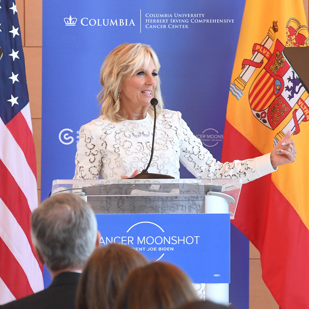 First Lady  Jill Biden speaks at podium in front of American and Spanish flags and the HICCC logo